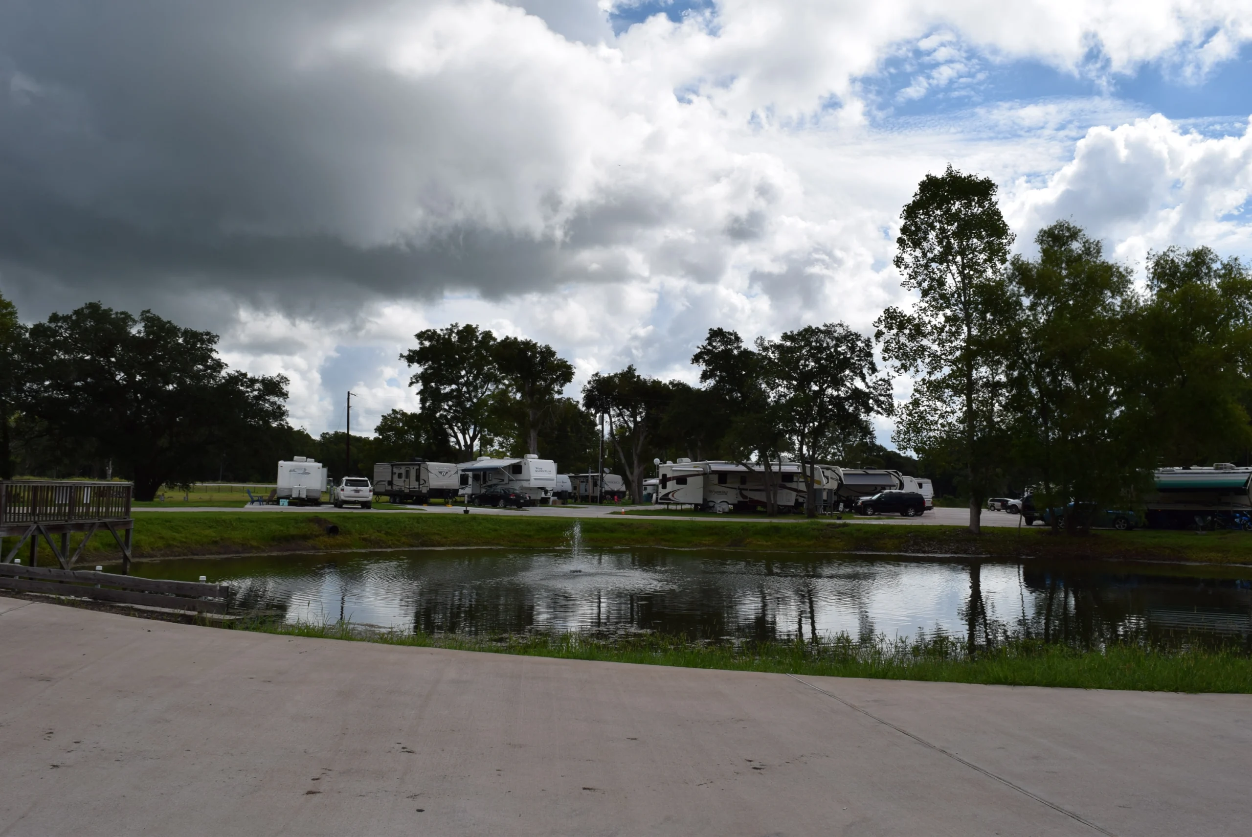 RVs and trucks parked along a tree-lined pond with a small fountain under a dramatic, partly cloudy sky at an RV park in Sweeny, Texas.