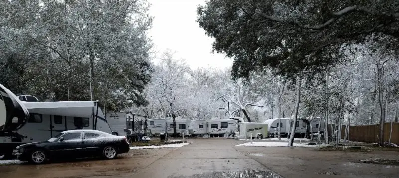 Snow-dusted trees line a quiet RV park road with trailers parked along both sides and a sedan in the foreground, showing winter conditions at a long-term stay site.